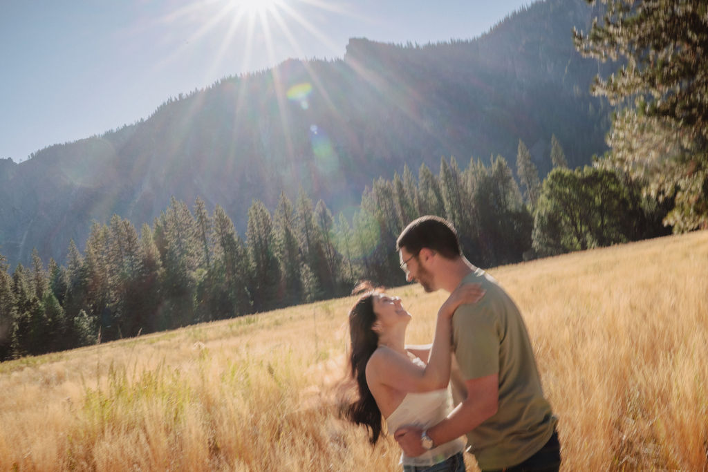 A couple smiling and embracing in front of a rocky river with tall mountains and trees in the background for their yosemite national park engagement photos