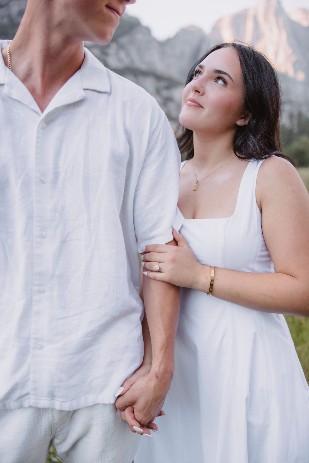 A couple in white attire stands in a grassy meadow with a backdrop of tall mountains and a waterfall.