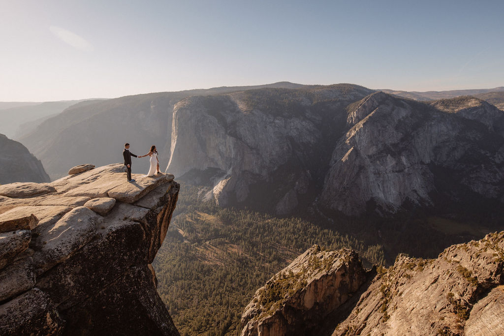 A couple dressed in wedding attire stands on a mountain ledge overlooking a vast rocky landscape with a prominent peak in the background. Best views in yosemite national park