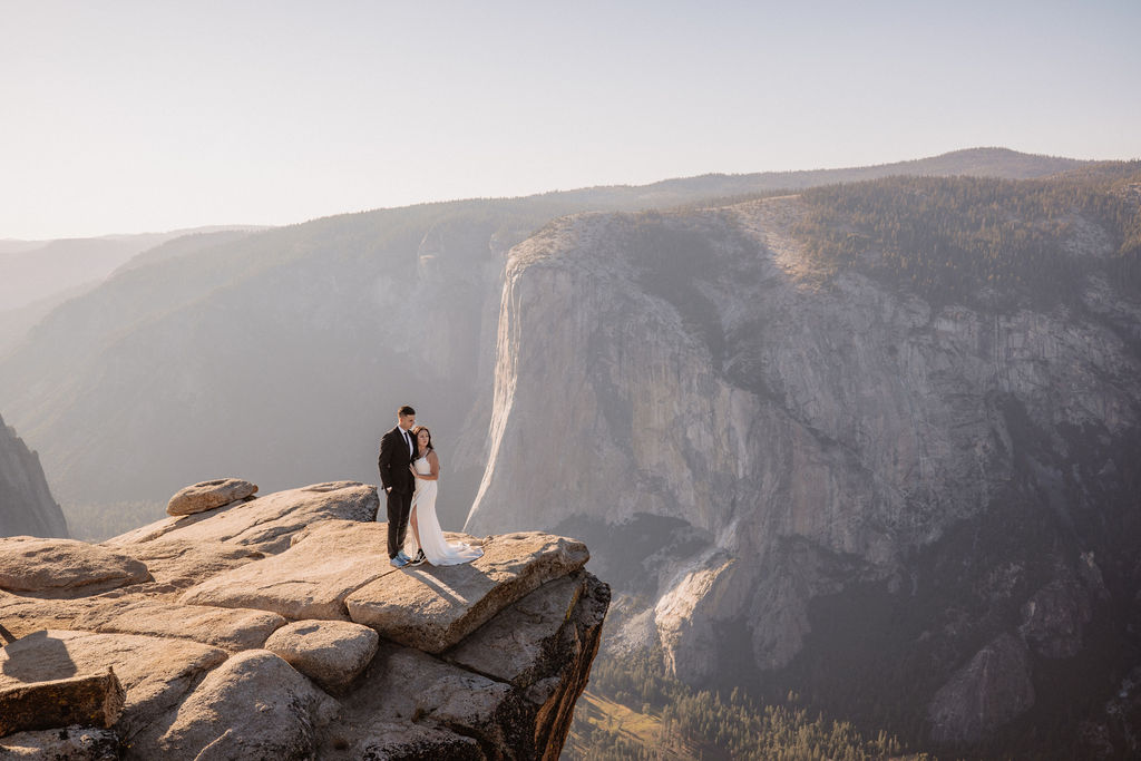 A couple dressed in wedding attire stands on a mountain ledge overlooking a vast rocky landscape with a prominent peak in the background. Best views in yosemite national park