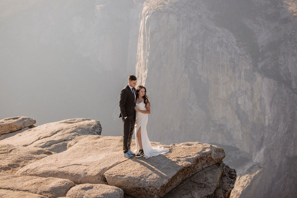 A couple dressed in wedding attire stands on a mountain ledge overlooking a vast rocky landscape with a prominent peak in the background. 