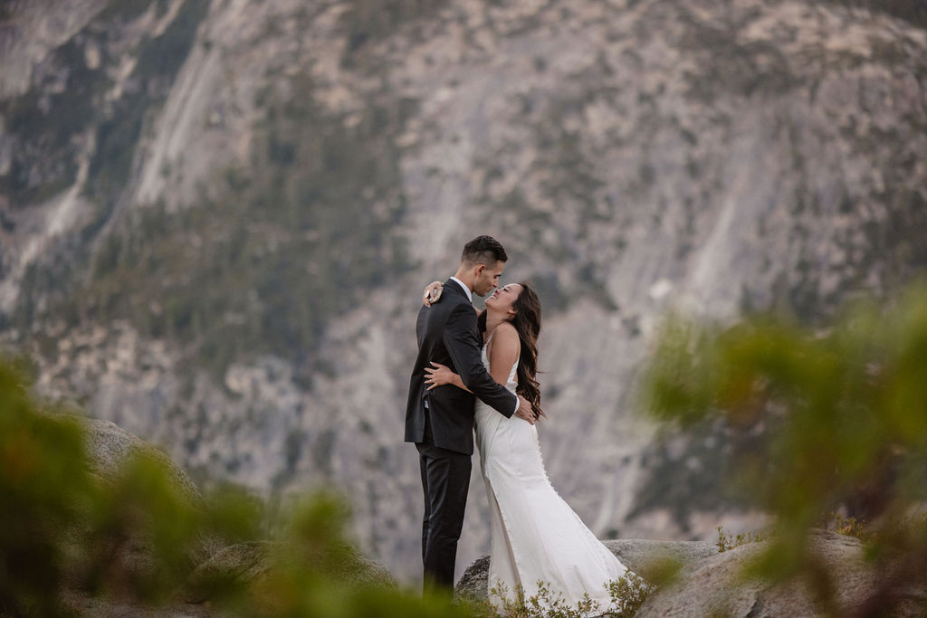 A couple dressed in wedding attire stands on a mountain ledge overlooking a vast rocky landscape with a prominent peak in the background. 