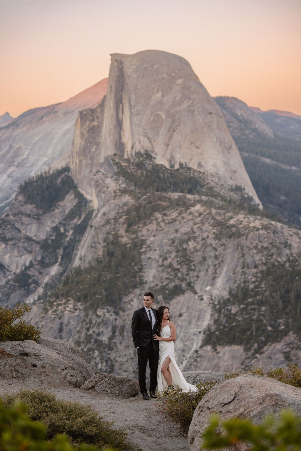 A couple dressed in wedding attire stands on a mountain ledge overlooking a vast rocky landscape with a prominent peak in the background. Best views in yosemite national park