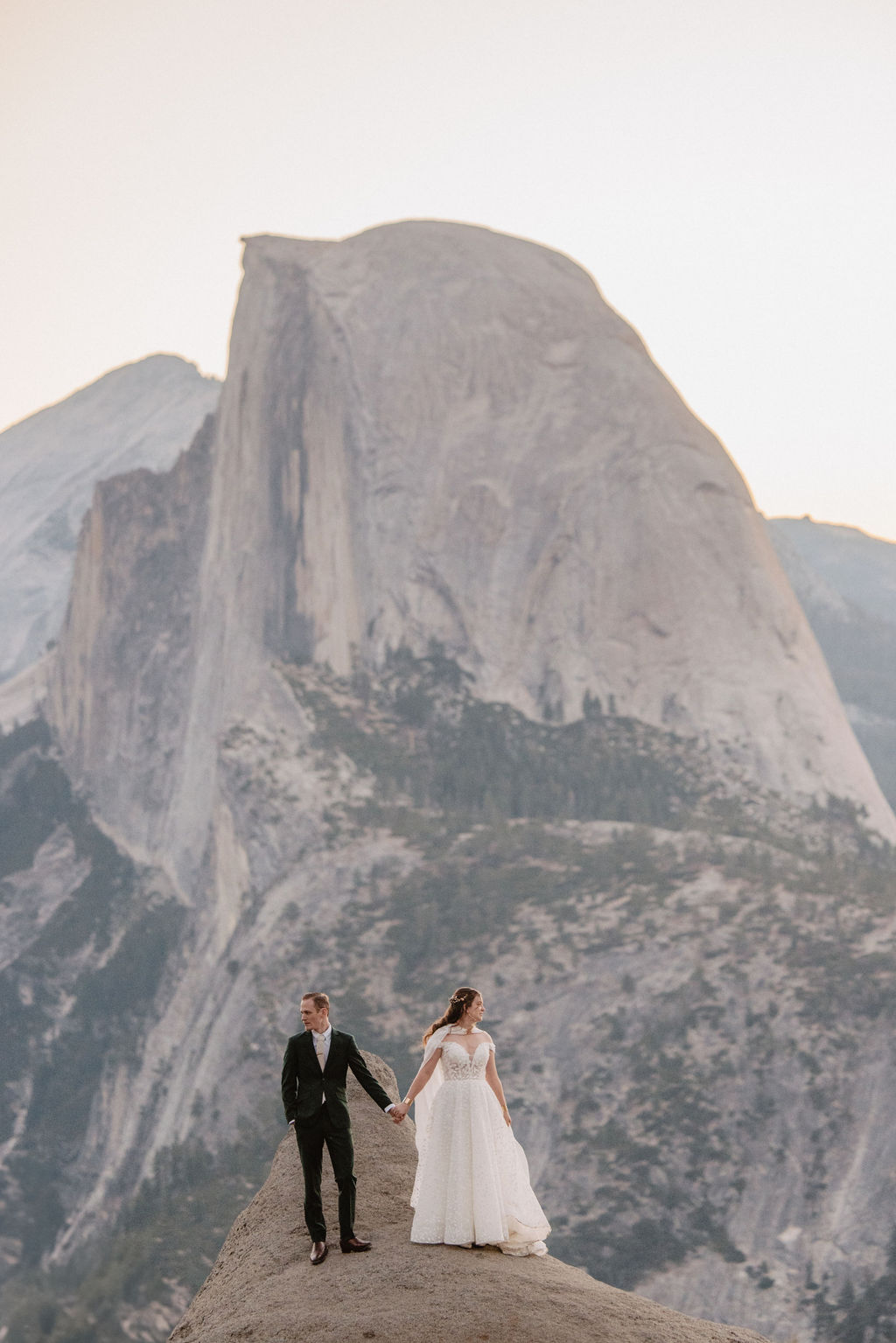 A couple in wedding attire stands on a rocky ledge, overlooking a mountainous landscape at sunrise at an elopement at Yosemite National park