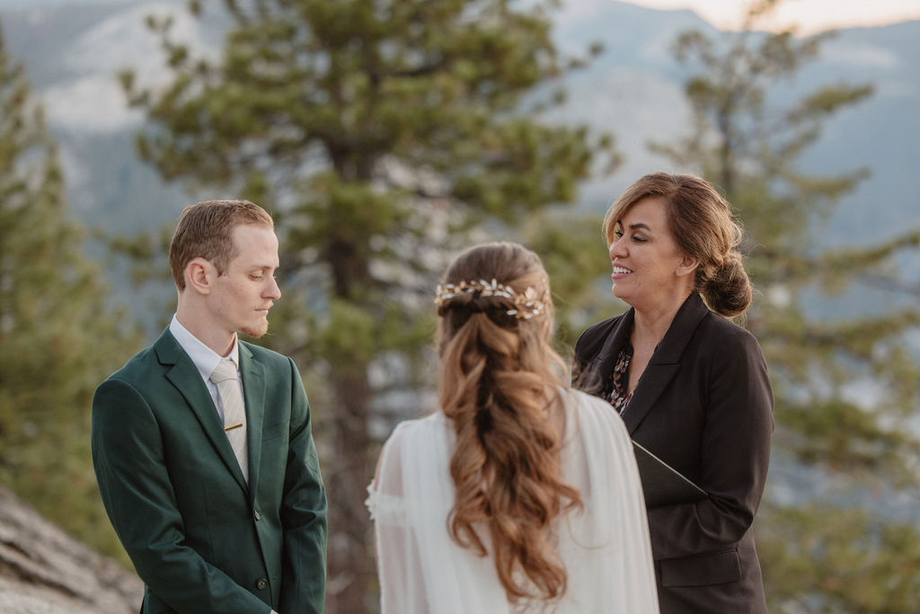 A couple exchanges vows with an officiant on a rocky overlook, with a mountain landscape in the background.