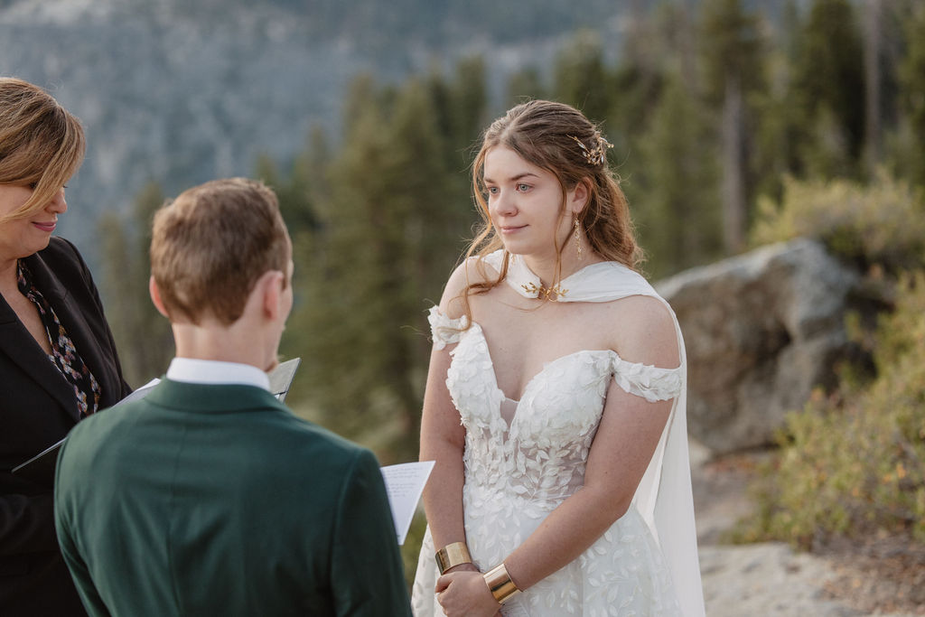 A couple exchanges vows with an officiant on a rocky overlook, with a mountain landscape in the background for a Yosemite elopement at Glacier Point and Mariposa Grove