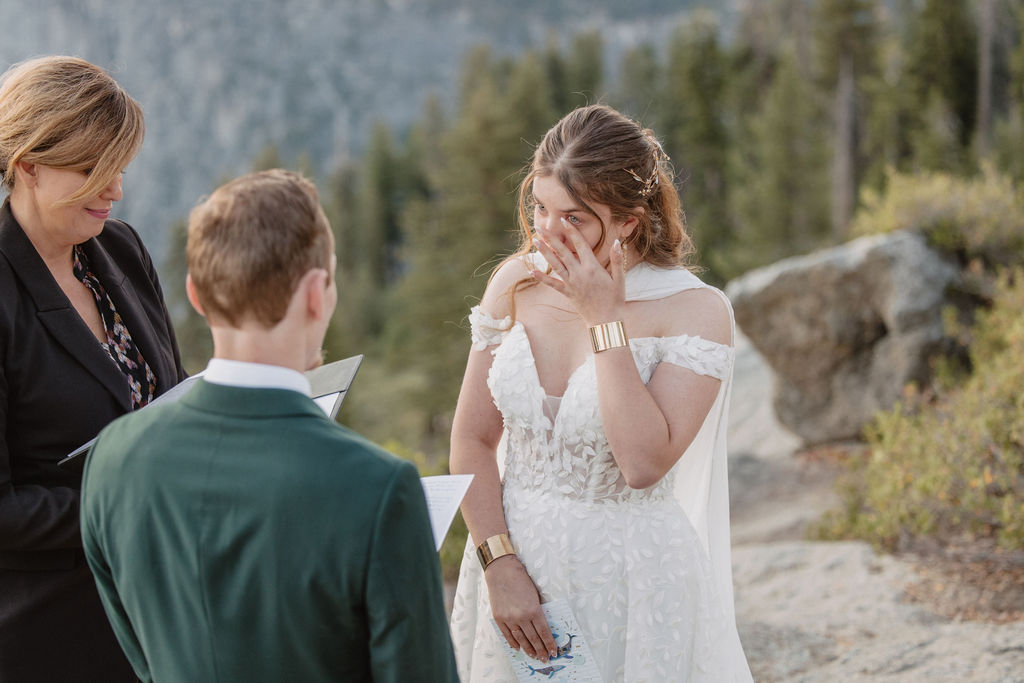 A couple exchanges vows with an officiant on a rocky overlook, with a mountain landscape in the background for a Yosemite elopement at Glacier Point and Mariposa Grove