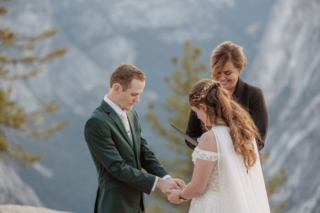 A couple exchanges vows with an officiant on a rocky overlook, with a mountain landscape in the background.