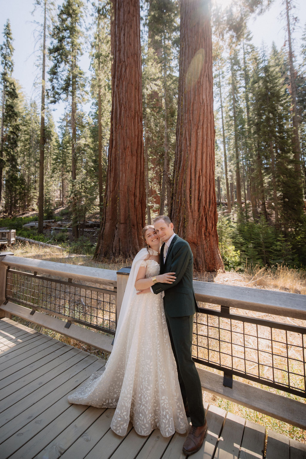 A couple in wedding attire walks through a forested path surrounded by tall trees at Mariposa Grove