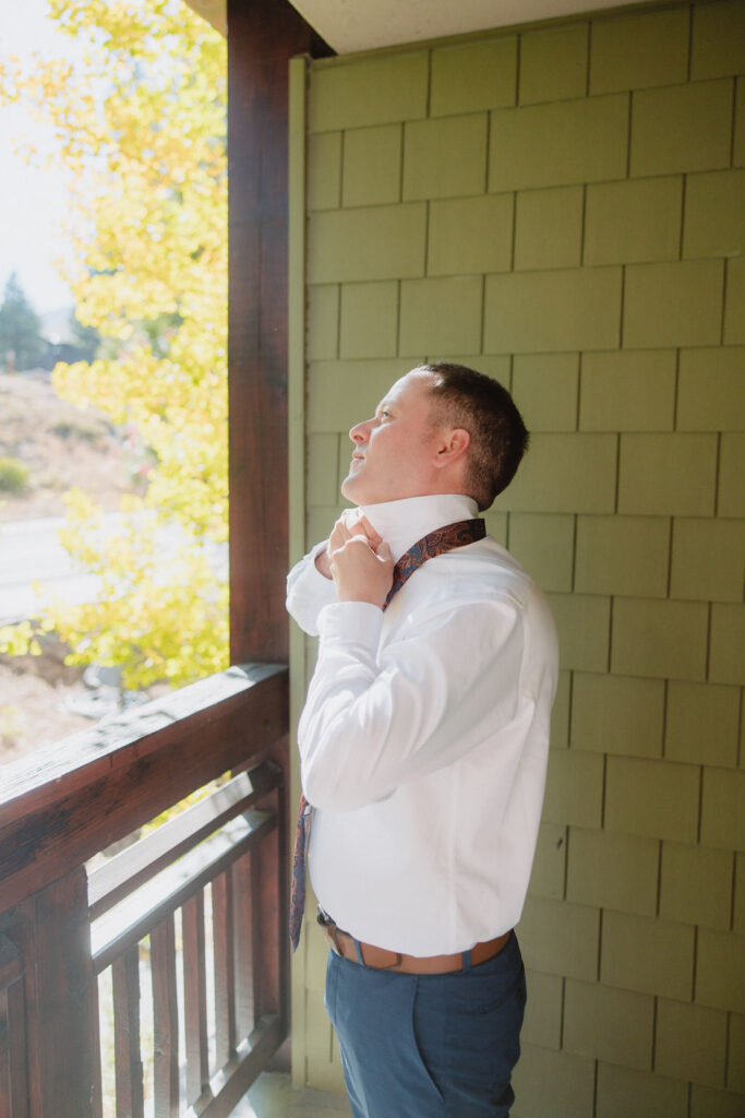 A person in a white shirt stands on a balcony, adjusting their tie. The background features green siding and autumn foliage for a wedding at sierra star golf course