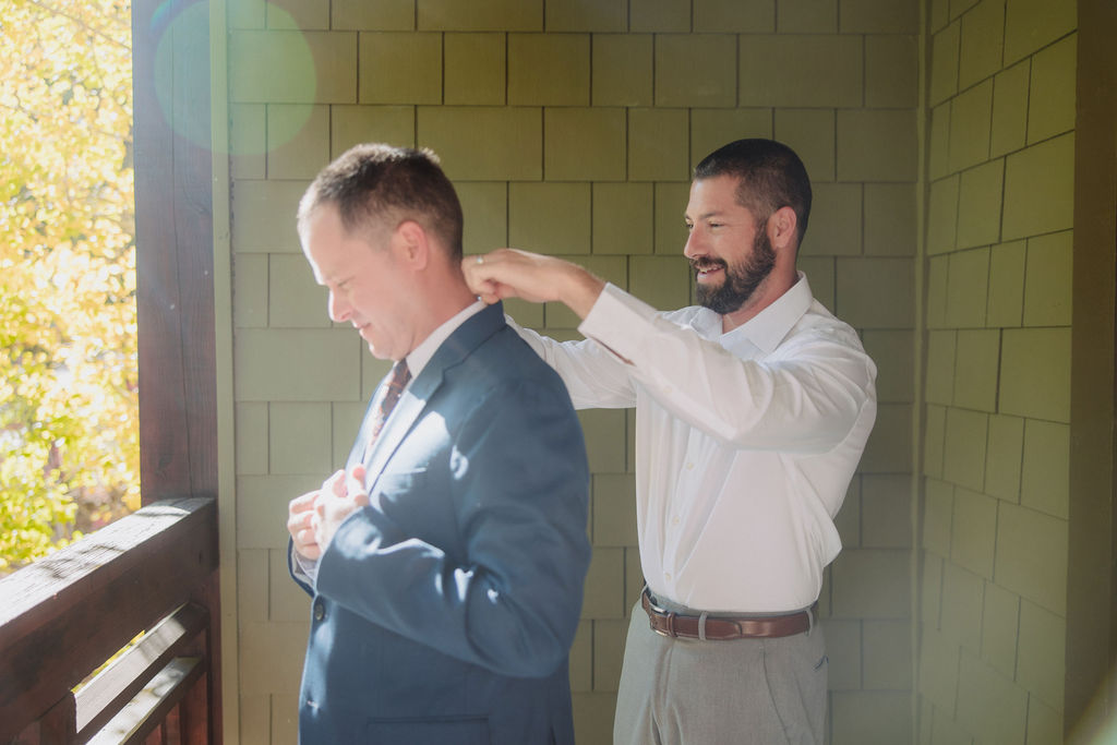 A person in a white shirt stands on a balcony, adjusting their tie. The background features green siding and autumn foliage for a wedding at sierra star golf course