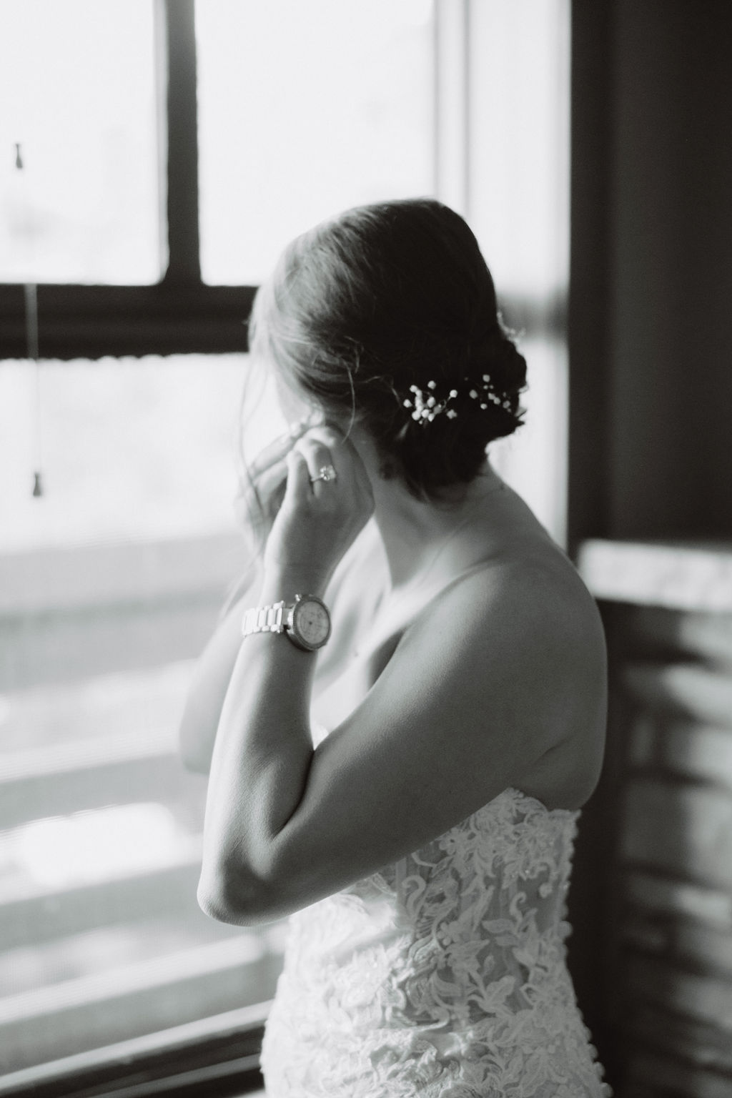 A woman in a white lace wedding gown stands facing a window beside a stone fireplace, with the gown's train spread out on the floor.