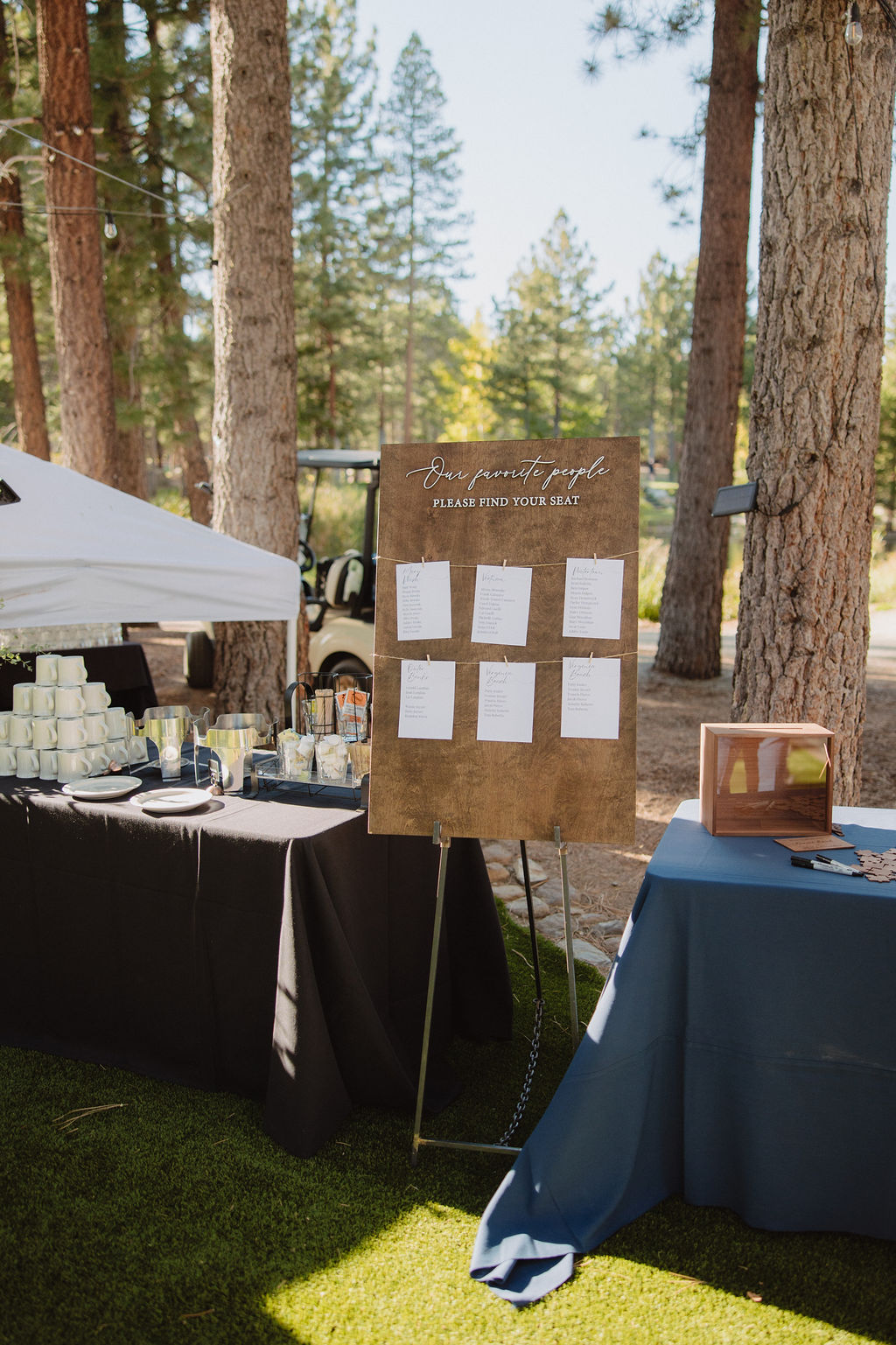 An outdoor event features a wooden board with paper notes, a table with stacked cups and drinks, and a blue cloth-covered table nearby, surrounded by tall trees.