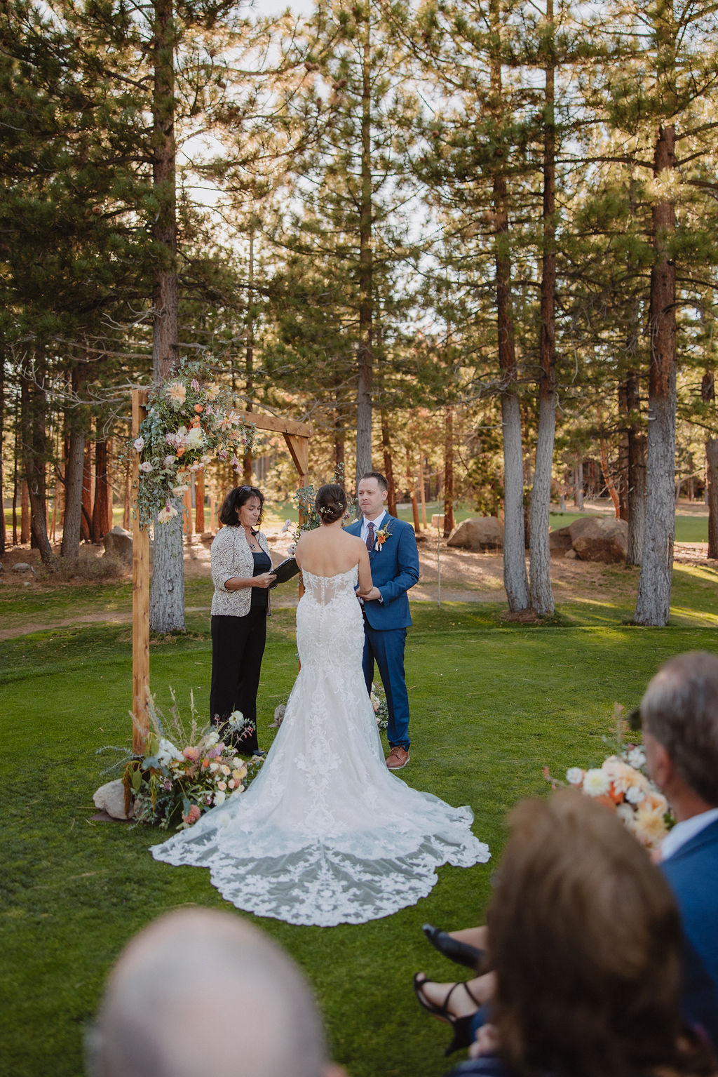 A couple stands at an outdoor wedding ceremony, facing the officiant, surrounded by trees and seated guests for their wedding at A Dreamy Sierra Star Golf Course Wedding in Mammoth