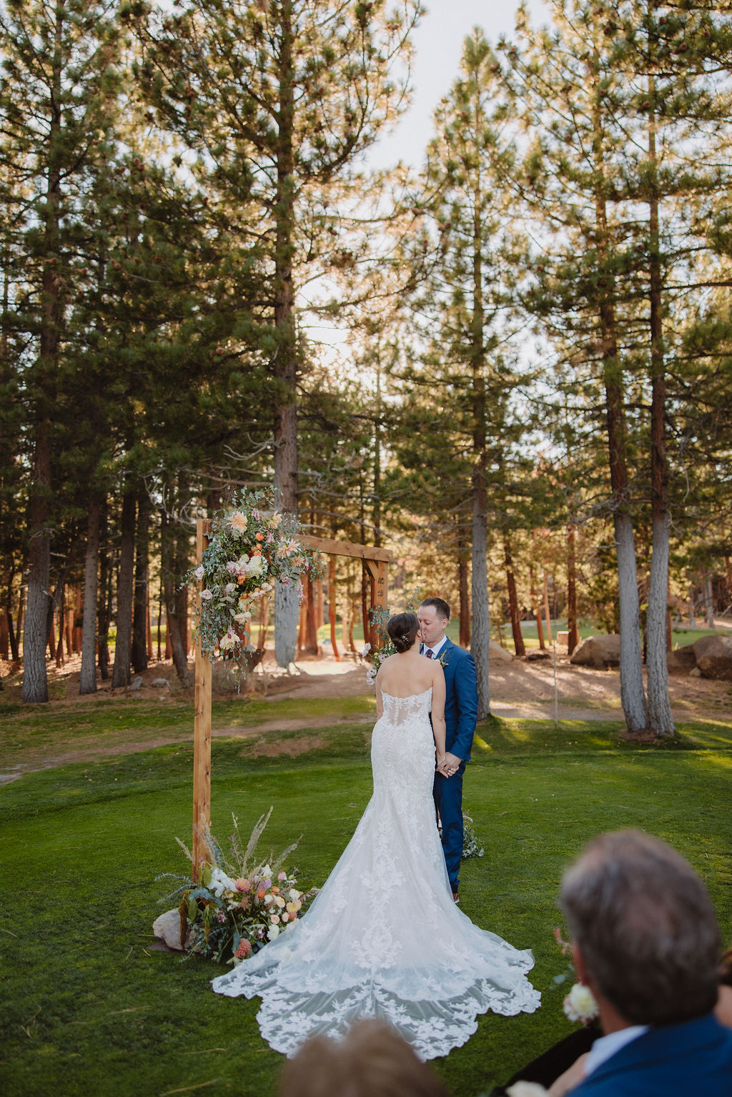 A couple stands at an outdoor wedding ceremony, facing the officiant, surrounded by trees and seated guests for their wedding at A Dreamy Sierra Star Golf Course Wedding in Mammoth