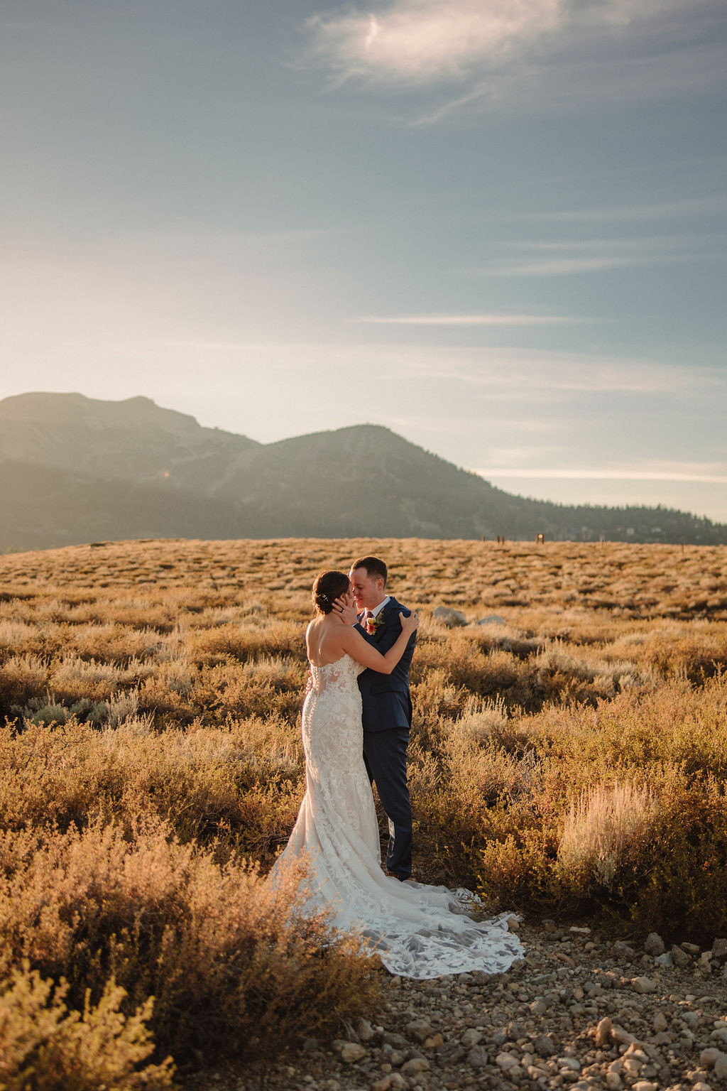 A couple embraces outdoors during sunset, with the sun shining in the background and mountains in the distance. The person in a wedding dress is seen from behindnfor their wedding at Sierra Star Golf Course