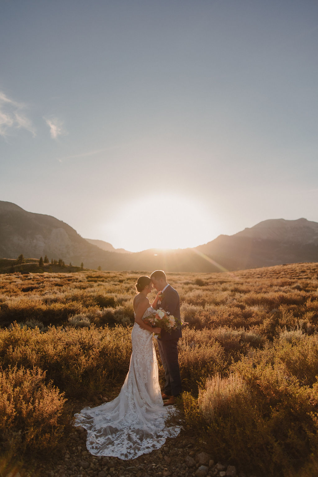 A couple embraces outdoors during sunset, with the sun shining in the background and mountains in the distance. The person in a wedding dress is seen from behindnfor their wedding at Sierra Star Golf Course