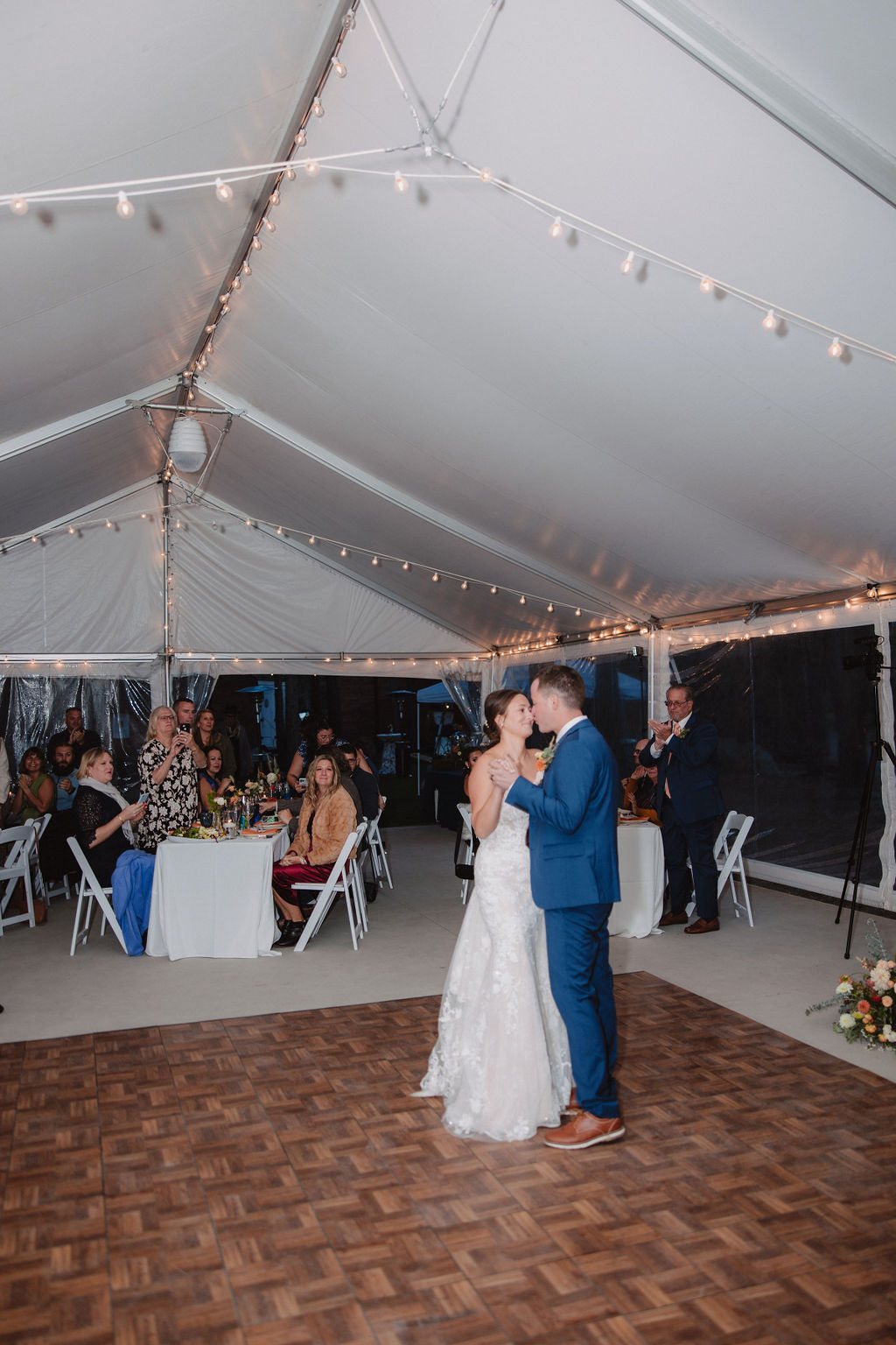 A couple dressed in wedding attire shares a dance under a white tent decorated with string lights, surrounded by seated guests at tables.