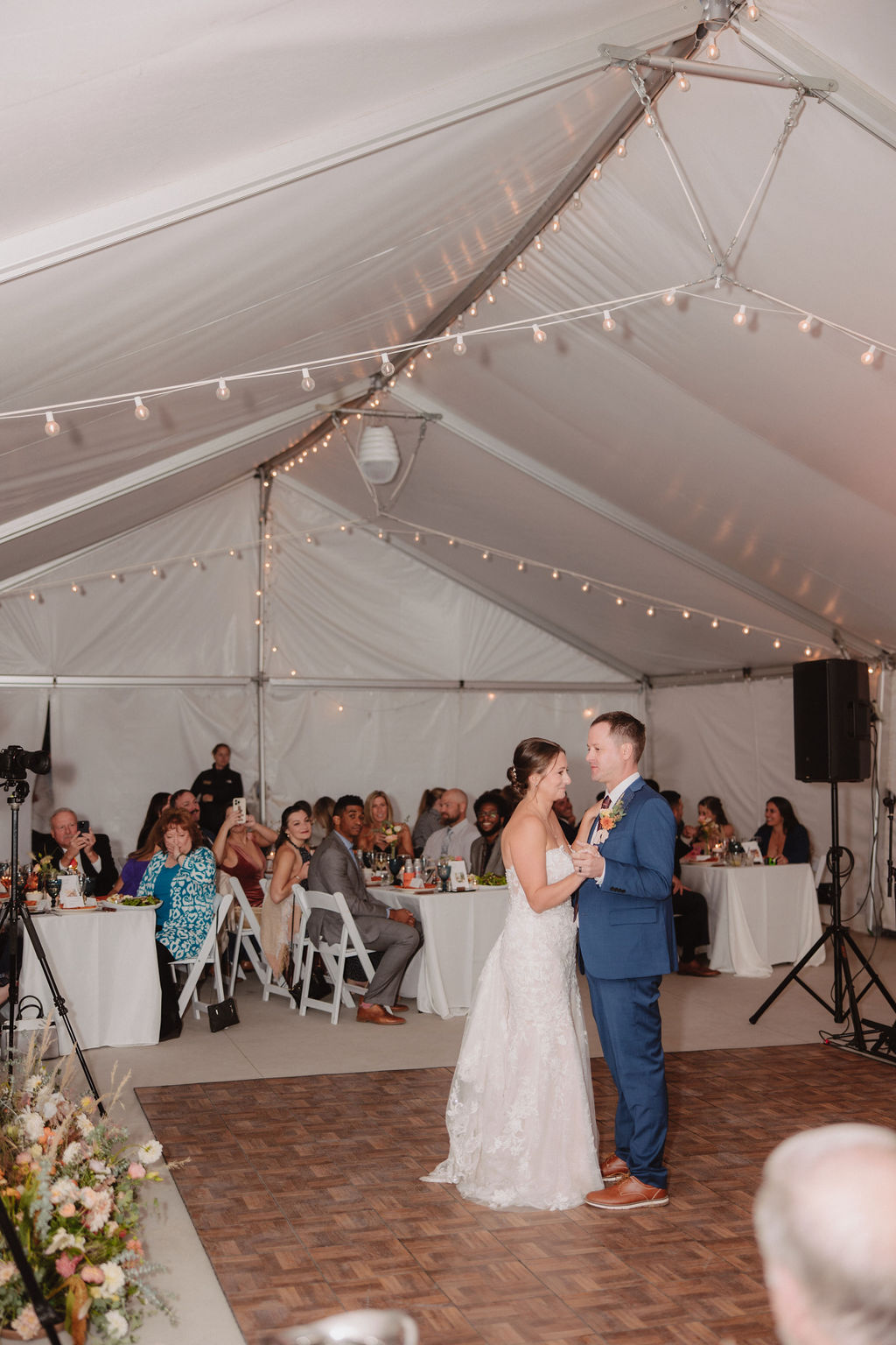 A couple dressed in wedding attire shares a dance under a white tent decorated with string lights, surrounded by seated guests at tables.