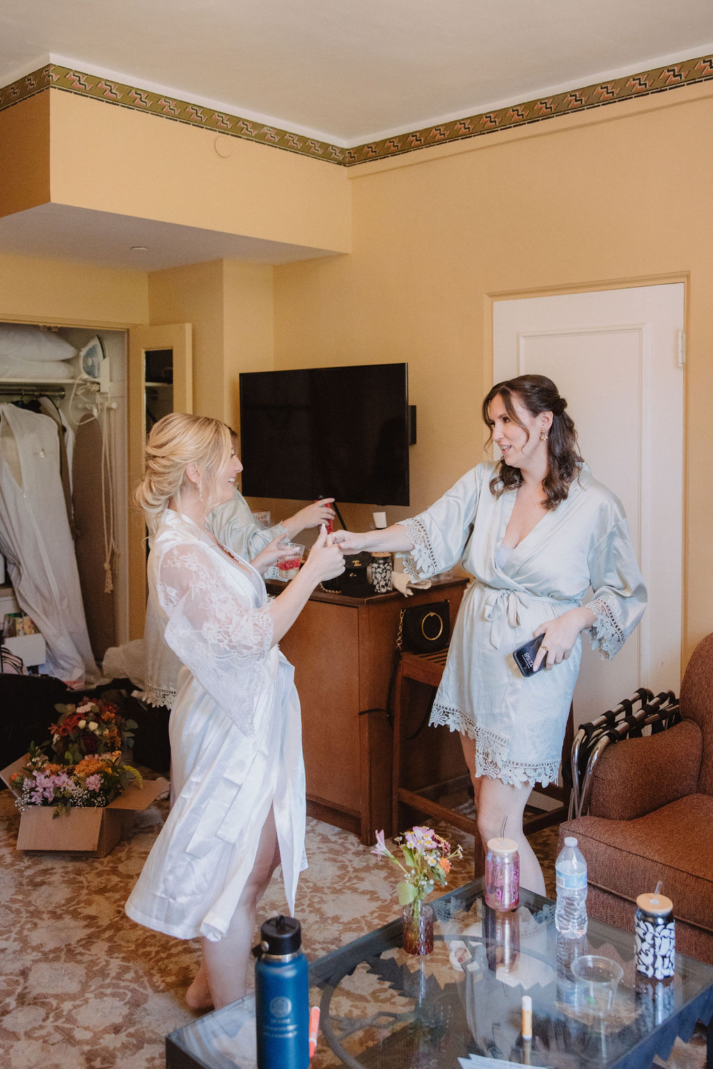 Two women in robes stand in a room, one holding a phone. The room has a TV, a table with drinks, and a closet with hanging clothes in the background.