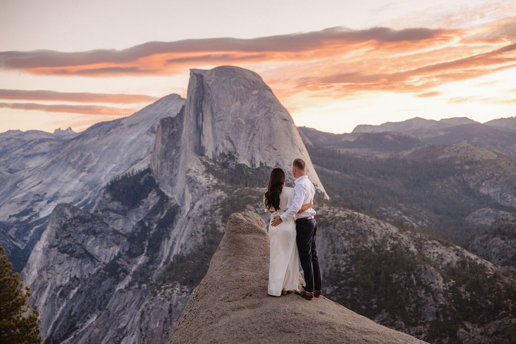 A couple stands on a rock ledge, gazing at Half Dome in Yosemite National Park, with a sunset sky in the background for their yosemite engagement photos