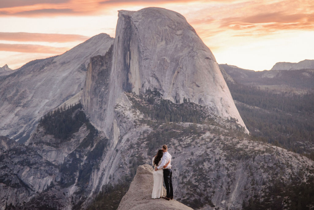 A couple stands on a rock ledge, gazing at Half Dome in Yosemite National Park, with a sunset sky in the background for their yosemite engagement photos