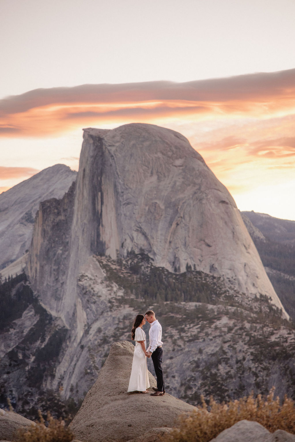 A couple stands on a rock ledge, gazing at Half Dome in Yosemite National Park, with a sunset sky in the background for their yosemite engagement photos