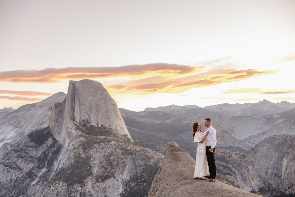 A couple stands on a rock ledge, gazing at Half Dome in Yosemite National Park, with a sunset sky in the background for their yosemite engagement photos