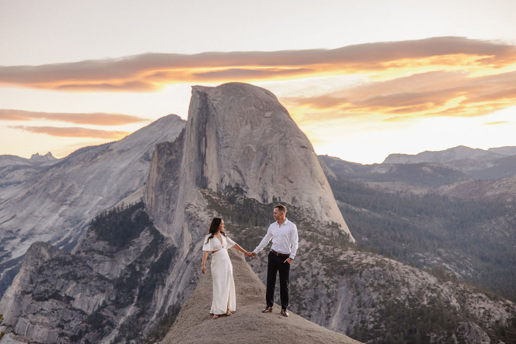 A couple stands on a rock ledge, gazing at Half Dome in Yosemite National Park, with a sunset sky in the background for their yosemite engagement photos