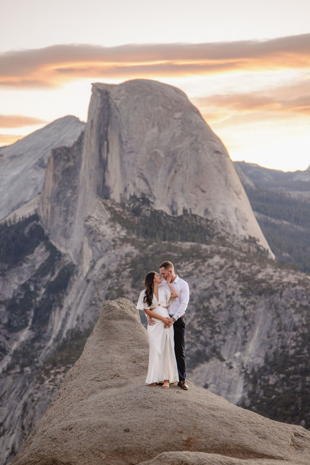 A couple stands on a rock ledge, gazing at Half Dome in Yosemite National Park, with a sunset sky in the background for their yosemite engagement photos