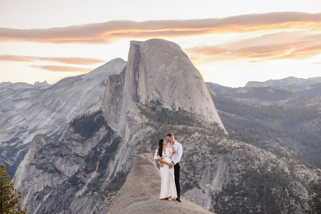 A couple stands on a rock ledge, gazing at Half Dome in Yosemite National Park, with a sunset sky in the background for their yosemite engagement photos