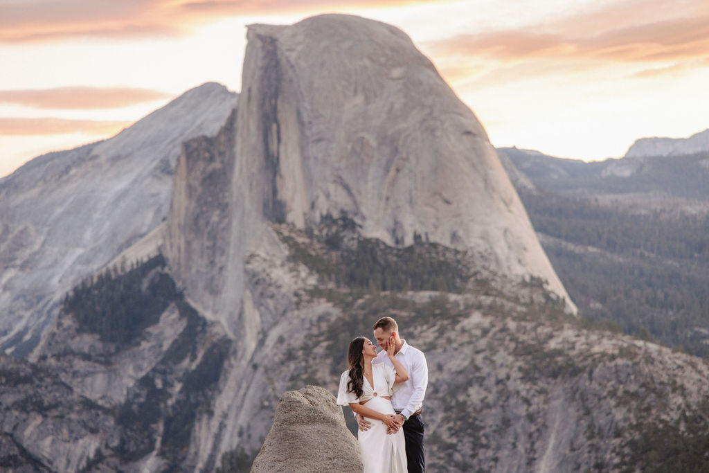 A couple embraces on a ledge with a large mountain, Half Dome, in the background at sunset