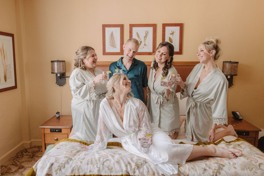 Five people in robes enjoy drinks on a bed in a warmly lit room with framed artwork on the wall.