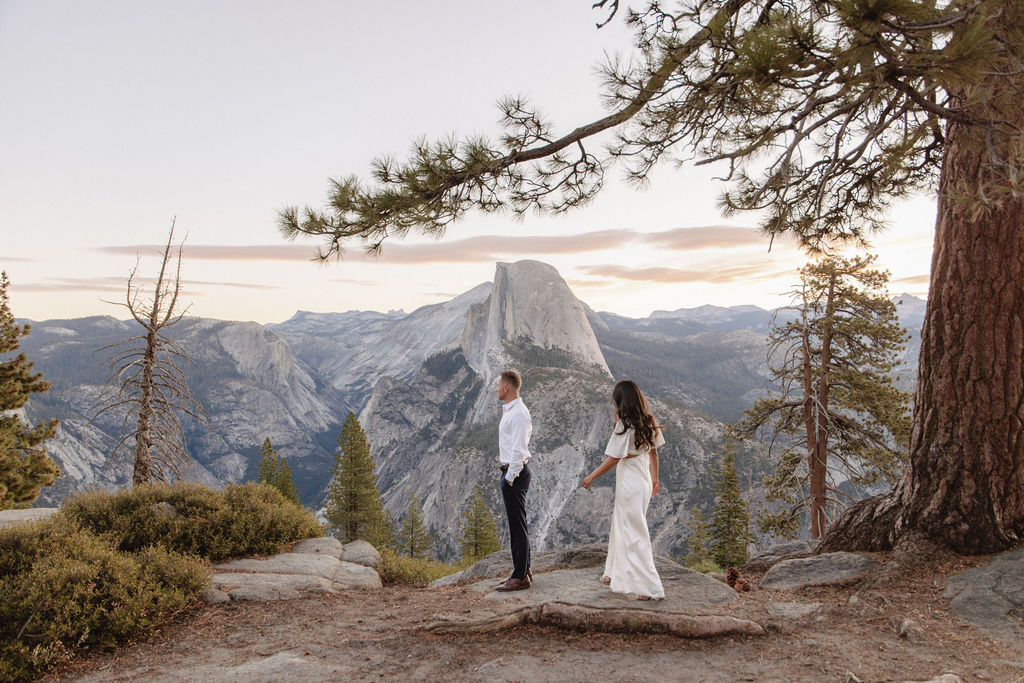 A couple stands on a rocky overlook with a view of a mountainous landscape, including a large rock formation, surrounded by trees.