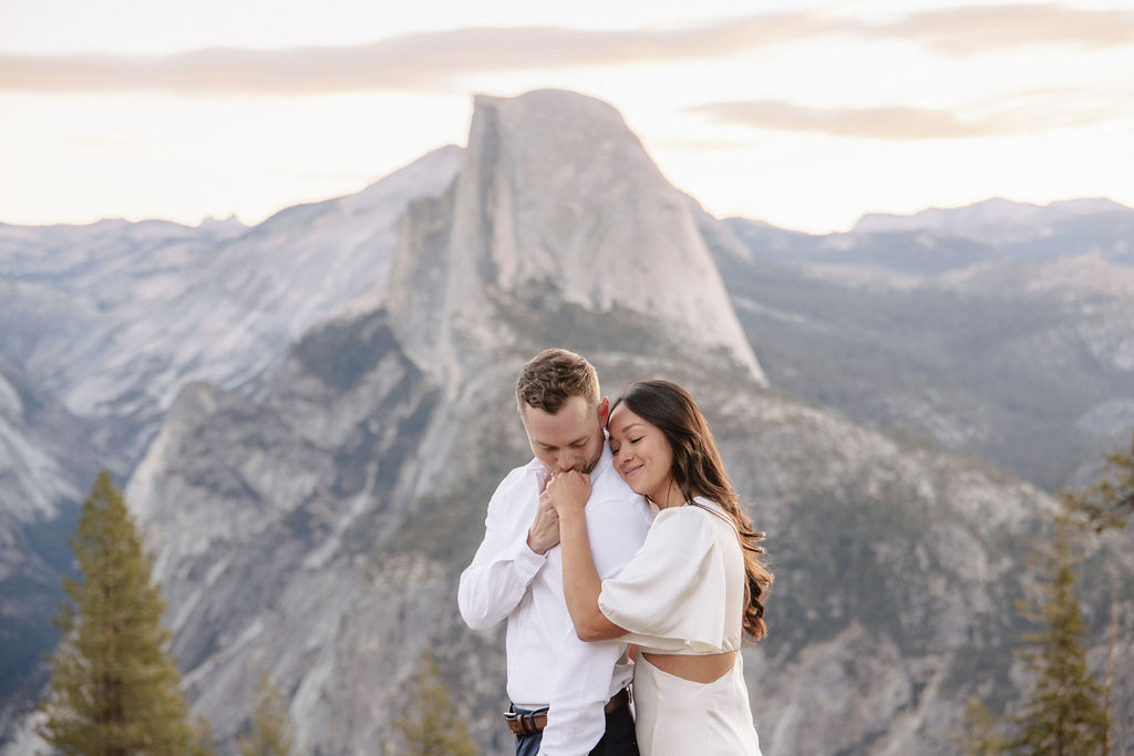 A couple stands on a rock ledge, gazing at Half Dome in Yosemite National Park, with a sunset sky in the background for their yosemite engagement photos