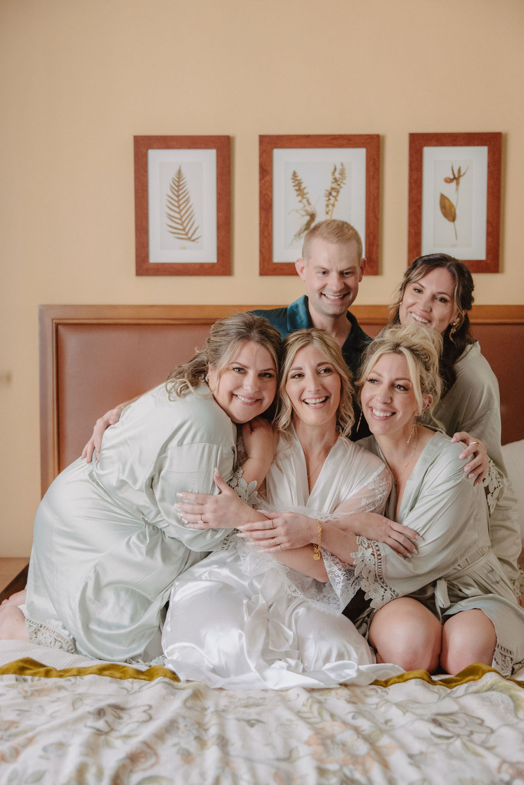 Five people in robes enjoy drinks on a bed in a warmly lit room with framed artwork on the wall.