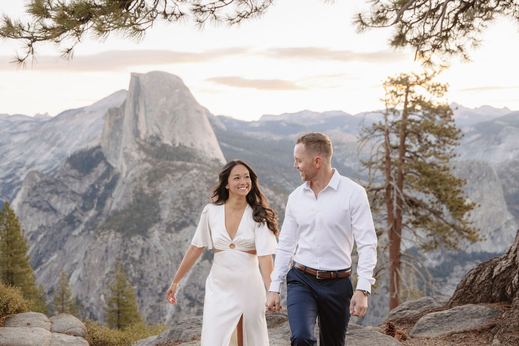 A couple stands on a rock ledge, gazing at Half Dome in Yosemite National Park, with a sunset sky in the background for their yosemite engagement photos