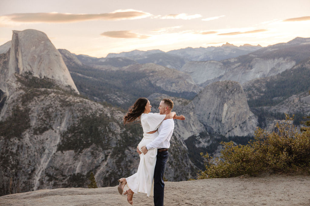 A couple embraces on a mountain cliff at sunset, with a scenic view of distant peaks and a partly cloudy sky.