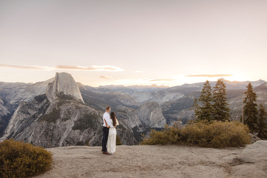 A couple stands on a rock ledge, gazing at Half Dome in Yosemite National Park, with a sunset sky in the background for their yosemite engagement photos