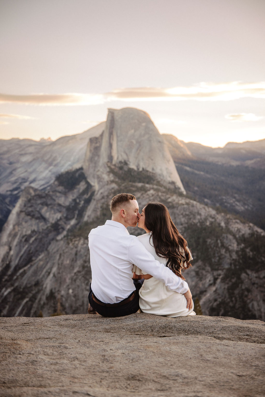 A couple stands on a rock ledge, gazing at Half Dome in Yosemite National Park, with a sunset sky in the background for their yosemite engagement photos