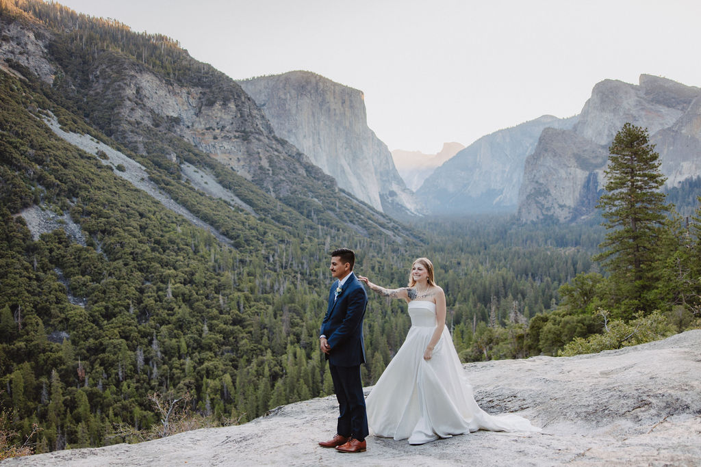 A bride and groom stand on a rocky ledge with a scenic view of a forested valley and mountains in the background for an elopement at bridalveil falls 