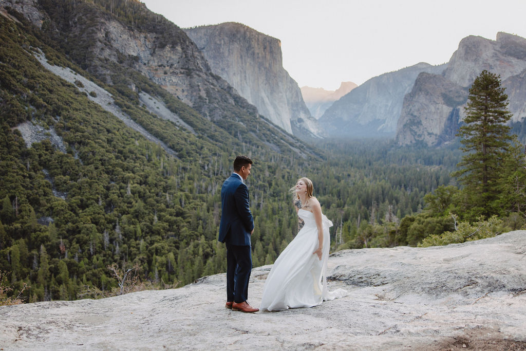 A bride and groom stand on a rocky ledge with a scenic view of a forested valley and mountains in the background for an elopement at bridalveil falls