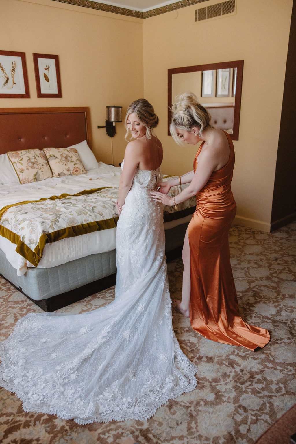 A woman in a white wedding dress stands in a bedroom while another woman in an orange dress helps adjust her gown.