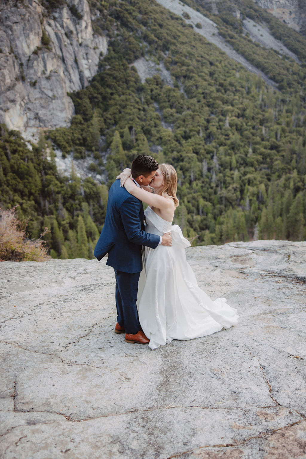 A bride and groom stand on a rocky ledge with a scenic view of a forested valley and mountains in the background for an elopement at bridalveil falls