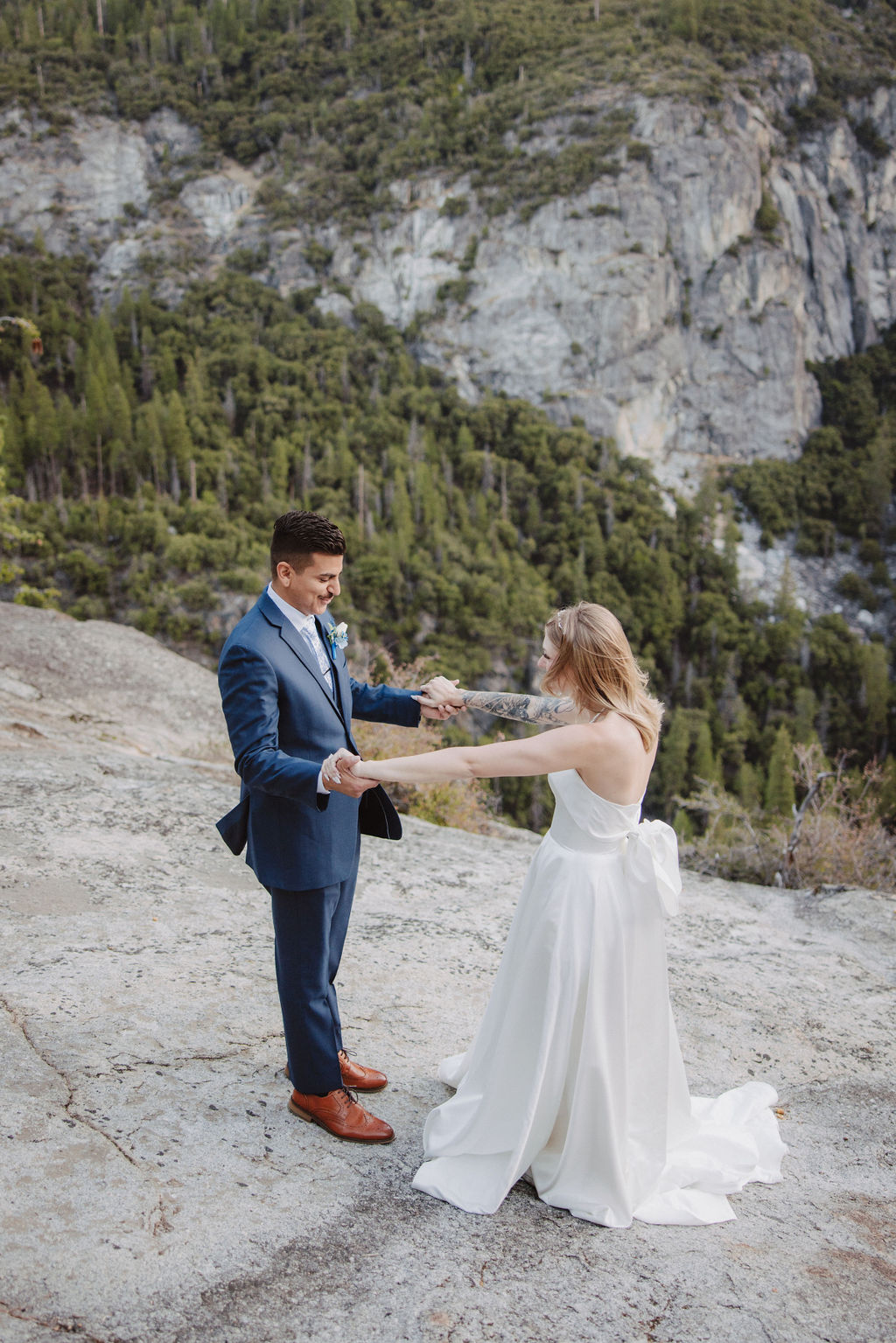 A bride and groom stand on a rocky ledge with a scenic view of a forested valley and mountains in the background for an elopement at bridalveil falls