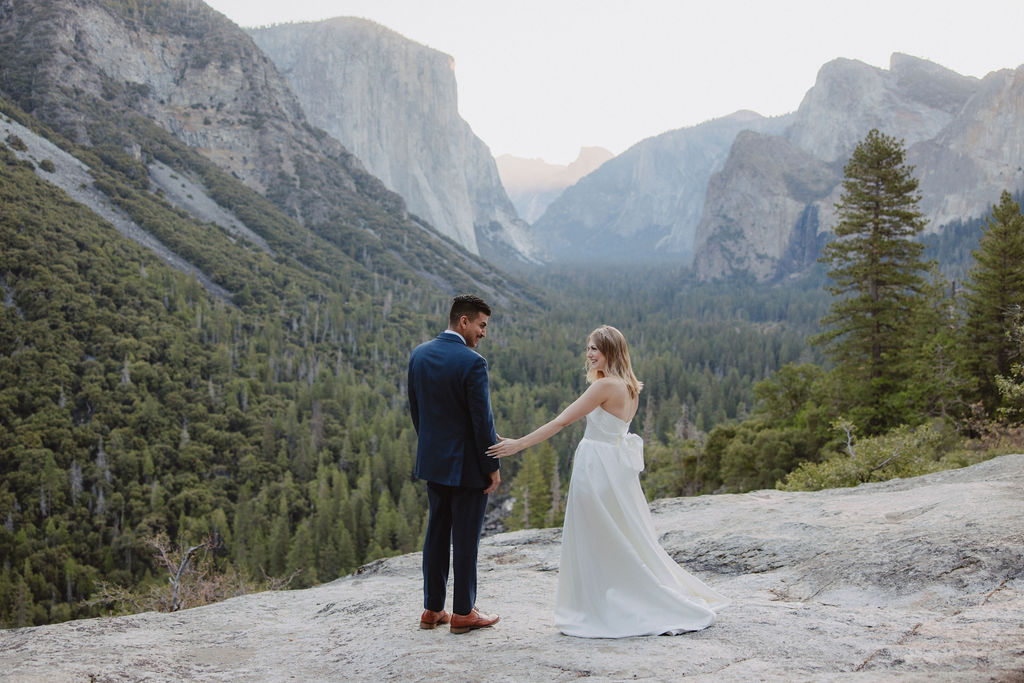 A bride and groom stand on a rocky ledge with a scenic view of a forested valley and mountains in the background for an elopement at bridalveil falls