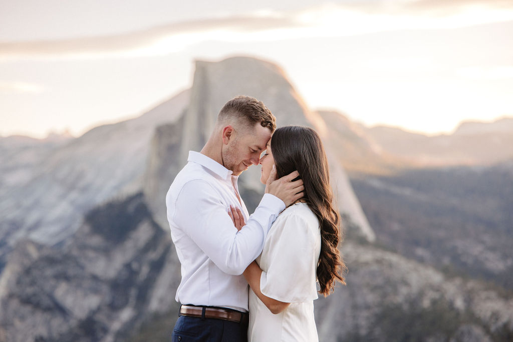 A couple stands on a rock ledge, gazing at Half Dome in Yosemite National Park, with a sunset sky in the background for their yosemite engagement photos