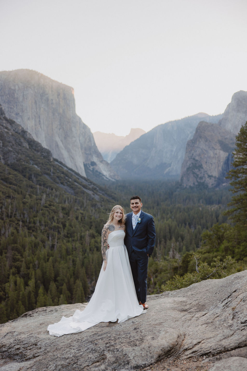 A bride and groom stand on a rocky ledge with a scenic view of a forested valley and mountains in the background for an elopement at bridalveil falls