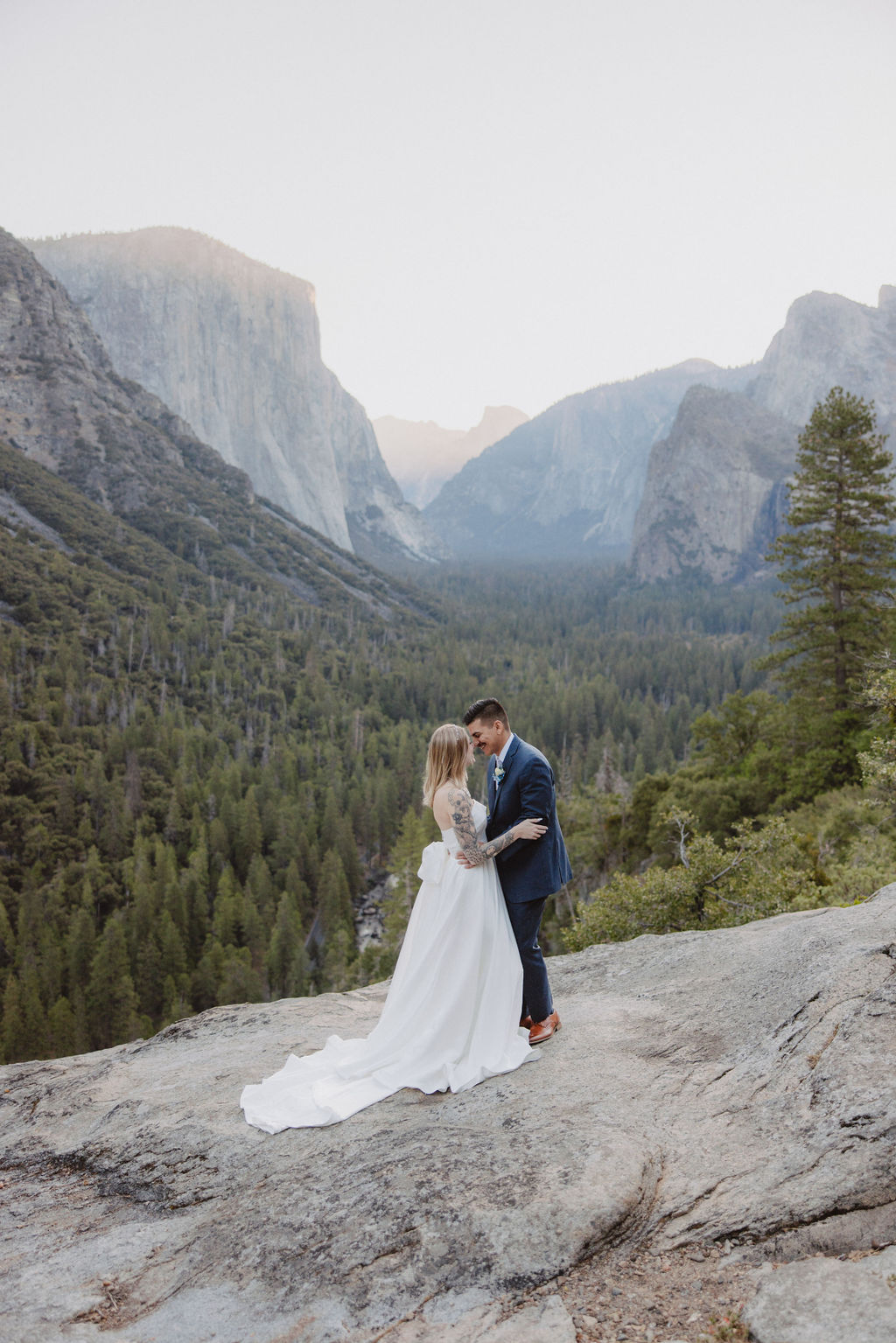 A bride and groom stand on a rocky cliff in wedding attire, with a forest and mountains in the background.