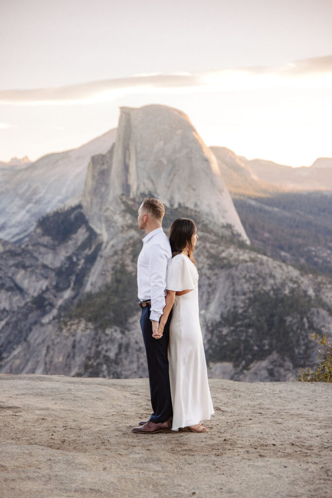 A couple stands on a rock ledge, gazing at Half Dome in Yosemite National Park, with a sunset sky in the background for their yosemite engagement photos